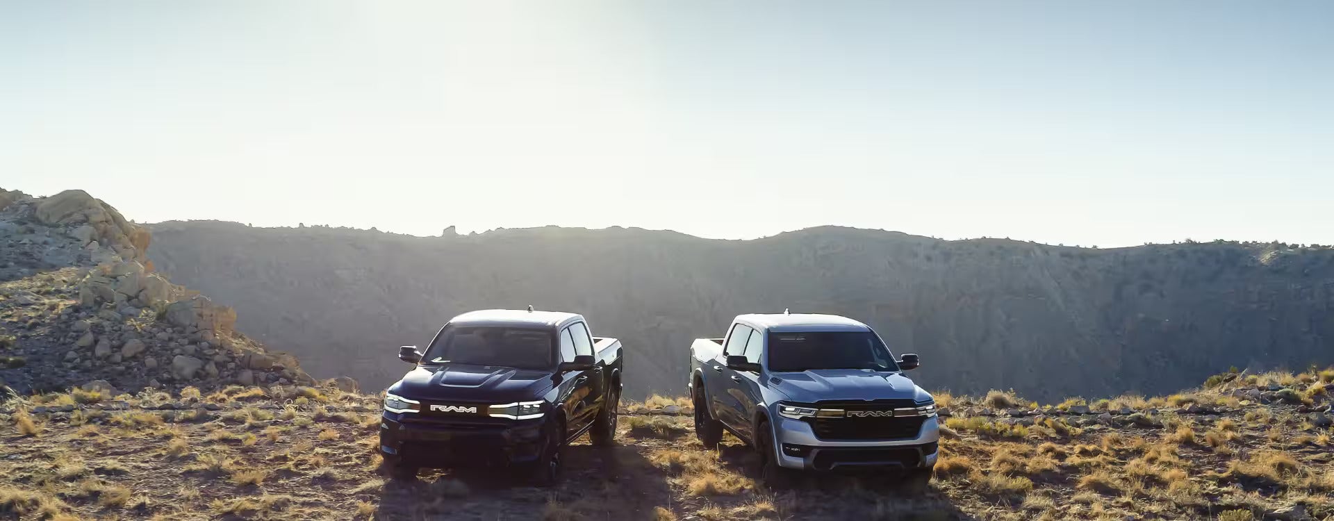 Two 2025 Ram 1500 REV trucks parked on a rocky terrain at sunrise, showcasing the vehicle’s off-road capability and rugged design.